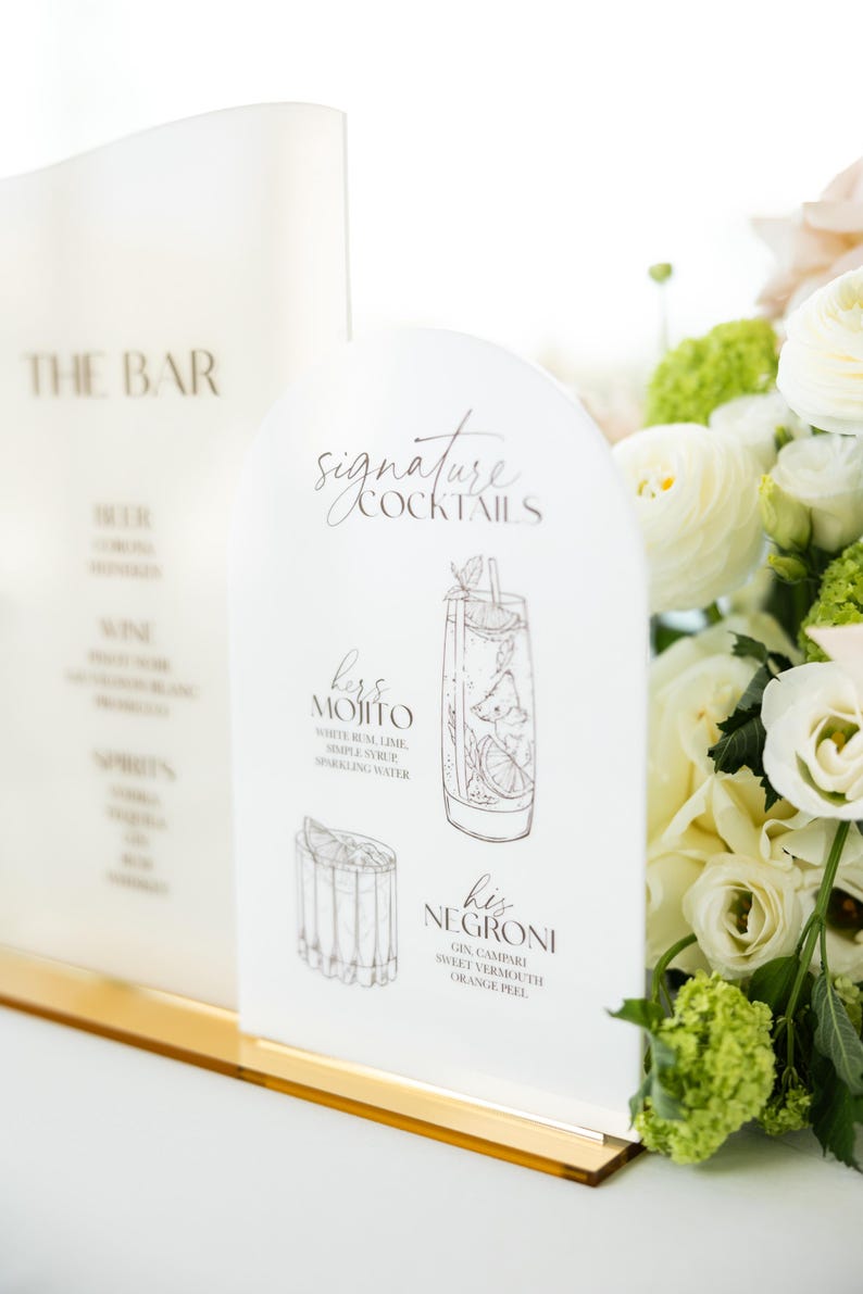 May include: White acrylic bar signs with gold trim. One sign reads "THE BAR" with a list of drinks. The other sign says "Signature Cocktails" and features illustrations of a Mojito and Negroni, with their ingredients.