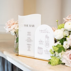 May include: White bar menu signs with gold accents, displaying "THE BAR" and "signature cocktails". The signs list drinks such as beer, wine, and spirits. The signs are decorated with flowers, and are suitable for a wedding.