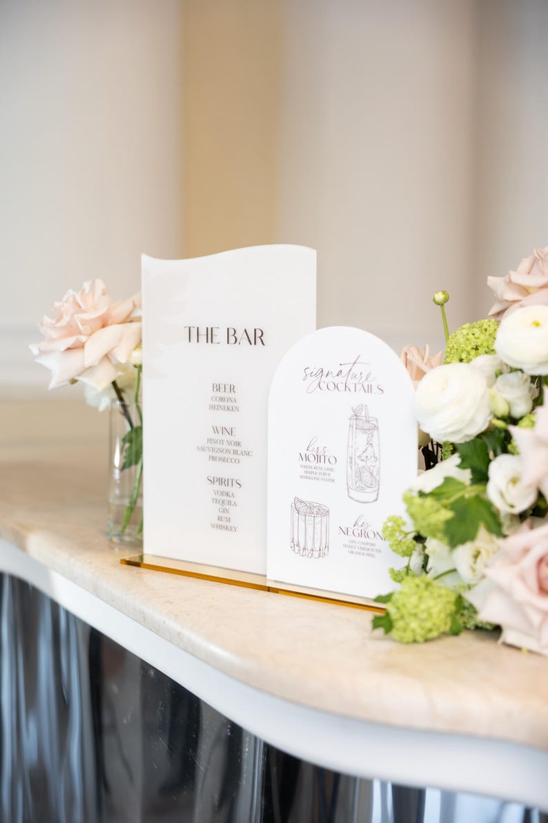 May include: White bar menu signs with gold accents, displaying "THE BAR" and "signature cocktails". The signs list drinks such as beer, wine, and spirits. The signs are decorated with flowers, and are suitable for a wedding.