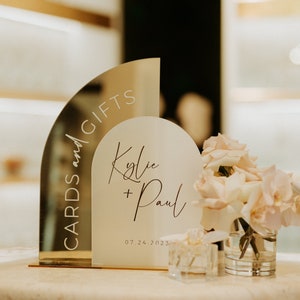 May include: A gold mirrored sign with the text "Cards and Gifts" and a white sign with the text "Kylie + Paul 07.24.2023". The signs are on a table with a vase of white roses.