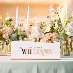 May include: A white table sign with gold lettering that reads "Mr. & Mrs. Williams". The sign is decorated with pink and white flowers in glass vases.