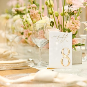 May include: A white card with gold lettering that says "Table 8" is placed on a table setting with white linens, clear glassware, and pink and white flowers.