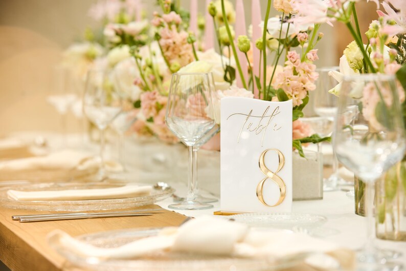 May include: A white card with gold lettering that says "Table 8" is placed on a table setting with white linens, clear glassware, and pink and white flowers.