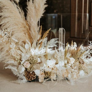 K&ouml;nnte beinhalten: Ein klares Acrylschild mit einem Blumengesteck. Das Arrangement besteht aus Pampasgras, Trockenblumen und Rosen in neutralen T&ouml;nen. Das Schild ist f&uuml;r Hochzeiten oder Veranstaltungen konzipiert.