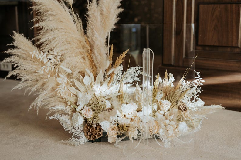 K&ouml;nnte beinhalten: Ein klares Acrylschild mit einem Blumengesteck. Das Arrangement besteht aus Pampasgras, Trockenblumen und Rosen in neutralen T&ouml;nen. Das Schild ist f&uuml;r Hochzeiten oder Veranstaltungen konzipiert.