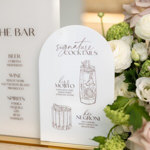 May include: White acrylic bar signs with elegant script. One sign lists drink categories: beer, wine, and spirits. The other features signature cocktails: a Mojito and a Negroni, with ingredients listed. Floral arrangement in the background.