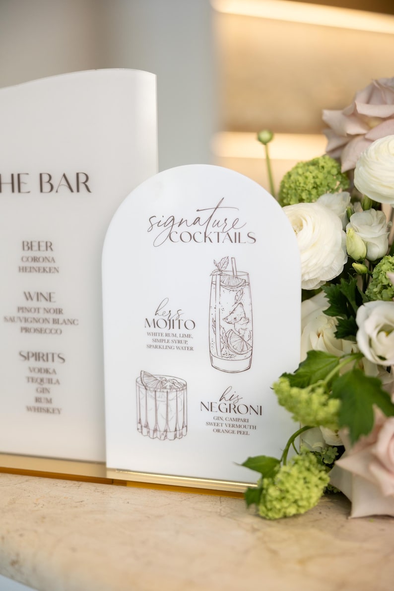May include: White acrylic bar signs with elegant script. One sign lists drink categories: beer, wine, and spirits. The other features signature cocktails: a Mojito and a Negroni, with ingredients listed. Floral arrangement in the background.
