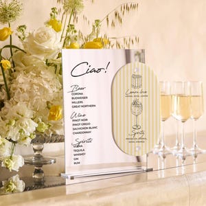 May include: A clear acrylic menu board with the word "Ciao!" and drink options, including beer, wine, and spirits. A smaller, striped sign features a cocktail illustration. Three champagne flutes are in the background, next to a floral arrangement.
