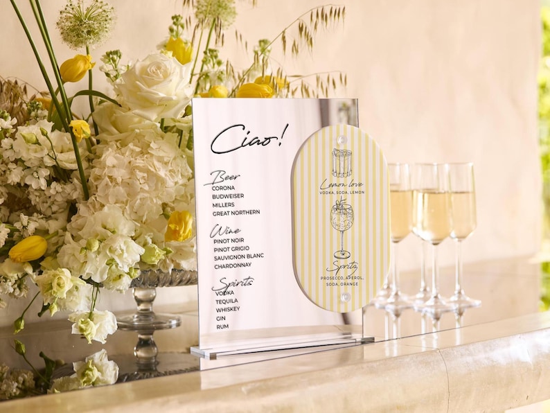 May include: A clear acrylic menu board with the word "Ciao!" and drink options, including beer, wine, and spirits. A smaller, striped sign features a cocktail illustration. Three champagne flutes are in the background, next to a floral arrangement.