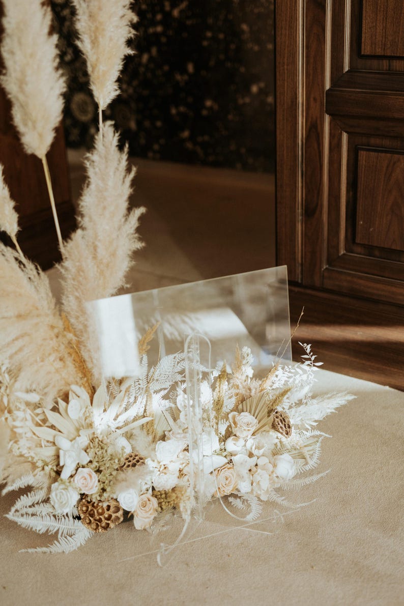 K&ouml;nnte beinhalten: Ein klares Acrylschild mit einem Blumenarrangement. Das Arrangement besteht aus wei&szlig;en und cremefarbenen Rosen, Trockenblumen und Pampasgras. Das Schild steht auf einem beigefarbenen Teppich, mit einer Holzt&uuml;r im Hintergrund.