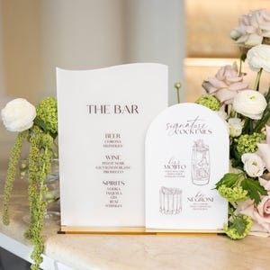 May include: Stylish bar menu signs displaying "THE BAR" and "Signature Cocktails". The menu lists beer, wine, and spirits. "Kiss Mojito" and "Her Negroni" are featured. Floral arrangements and a marble countertop are also visible.