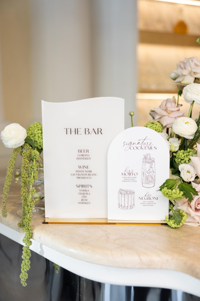 May include: Stylish bar menu signs displaying "THE BAR" and "Signature Cocktails". The menu lists beer, wine, and spirits. "Kiss Mojito" and "Her Negroni" are featured. Floral arrangements and a marble countertop are also visible.