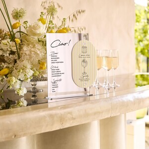 May include: A bar scene with a floral arrangement of white and yellow flowers, a menu with the word "Ciao!" and a list of drinks, and three champagne flutes filled with a golden liquid. The bar has a light-colored marble countertop.