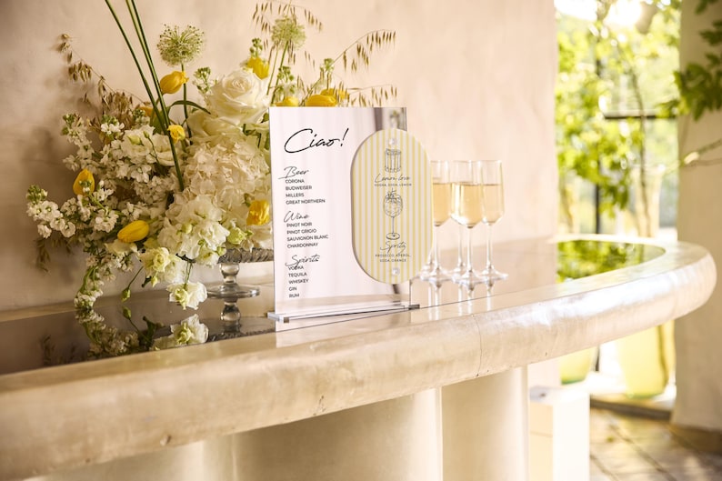 May include: A bar scene with a floral arrangement of white and yellow flowers, a menu with the word "Ciao!" and a list of drinks, and three champagne flutes filled with a golden liquid. The bar has a light-colored marble countertop.