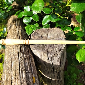 Op de afbeelding: Een houten toverstaf met een lichtbruine kleur en een donkerbruine band bij de handgreep. De toverstaf ligt op een houten boomstam.