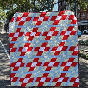 Pode incluir: Uma colcha retangular com um padrão geométrico repetitivo de quadrados vermelhos, brancos e verde-pálidos dispostos em grade. O design é atravessado por faixas diagonais de tecido azul-claro, criando uma superfície têxtil estruturada.