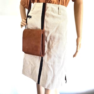 Könnte beinhalten: Eine braune Leder-Umhängetasche mit Klappenverschluss. Die Tasche wird über einem beigen Rock mit einem schwarzen Streifen an der Seite getragen. Die Schaufensterpuppe trägt ein braunes Oberteil.