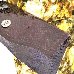 Könnte beinhalten: Eine dunkelbraune Clutch mit einem strukturierten, krokodilartigen Muster. Die Clutch hat einen Klappenverschluss und einen kleinen, runden silberfarbenen Knopf mit einem verzierten Design. Der Hintergrund zeigt ein Blumenmuster in Gelb und Grün.