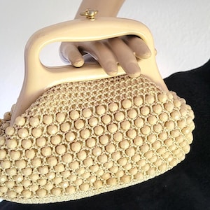 Könnte beinhalten: Eine beige Handtasche mit einem strukturierten, glatten, hellen Tragegriff und einem kleinen goldfarbenen Verschluss. Der Körper der Tasche ist mit einem dichten, gleichmäßigen Muster aus kleinen, runden, beigen Perlen in einer gewebten Gitterstruktur bedeckt.