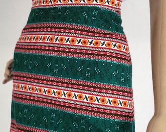 May include: A high-waisted midi skirt featuring a deep green base with repeating horizontal bands of orange floral patterns, white geometric lines, and small white dot motifs. The skirt is paired with a plain white short-sleeved t-shirt.