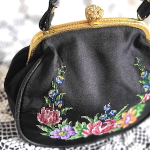 Könnte beinhalten: Schwarze Vintage-Handtasche mit goldfarbenem Rahmen und Griff. Die Tasche hat eine farbenfrohe Blumenstickerei in Rot-, Lila-, Blau- und Orangetönen. Der Verschluss ist mit einer dekorativen goldfarbenen Verzierung versehen.