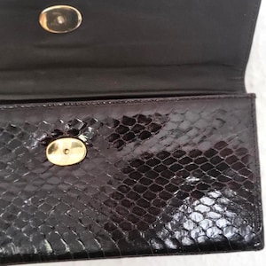 Pode incluir: Clutch preta com padrão de pele de cobra. A clutch tem um fecho dourado e um interior preto. A aba está aberta, revelando o interior da bolsa. A clutch é retangular.