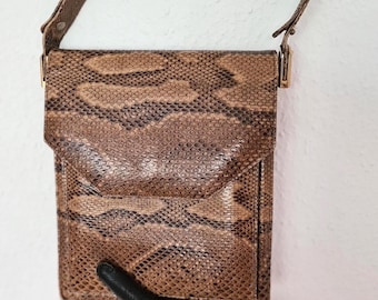 May include: A rectangular shoulder bag featuring a brown and tan snakeskin pattern. The bag has a fold-over flap closure, a matching snakeskin shoulder strap, and gold-toned metal hardware attachments connecting the strap to the main body.
