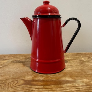 May include: A red enamel coffee pot with a black handle and rim. The pot has a lid and a spout for pouring. The coffee pot is sitting on a wooden surface.