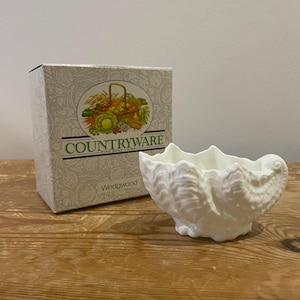 May include: A white ceramic shell-shaped dish with detailed design, beside a box with "COUNTRYWARE" and "Wedgwood" text. The dish sits on a wooden surface.