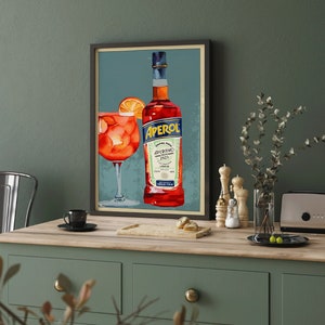 K&ouml;nnte beinhalten: Ein gerahmtes Vintage-Poster mit einer Flasche Aperol Aperitivo Lik&ouml;r und einem Glas Aperol Spritz. Das Poster hat einen blauen Hintergrund mit einem roten und wei&szlig;en Etikett auf der Flasche.