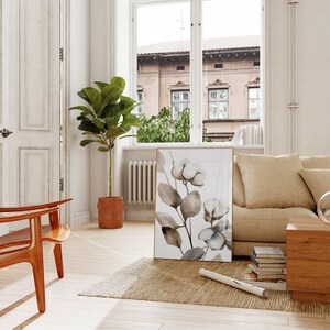 May include: A watercolor painting of a branch of cotton blossoms with brown leaves on a white background. The painting is framed in a gold frame and is hanging on a white wall in a living room.