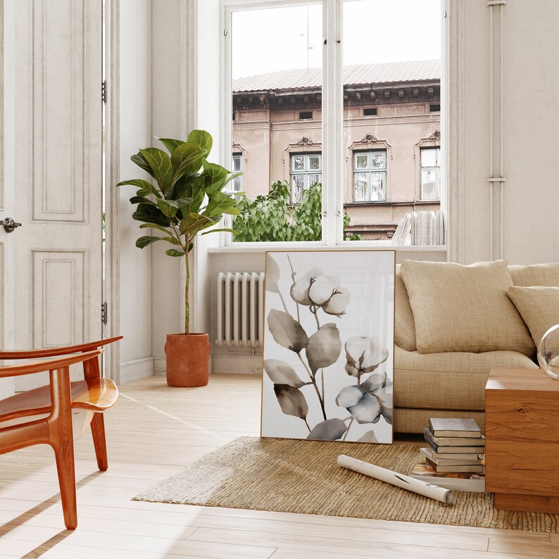 May include: A watercolor painting of a branch of cotton blossoms with brown leaves on a white background. The painting is framed in a gold frame and is hanging on a white wall in a living room.