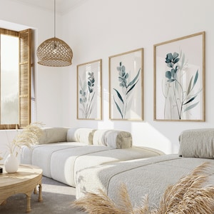 May include: Three framed botanical prints with a light background and blue-green foliage designs hang on a white wall above a white couch with two pillows. The couch is in a living room with a woven light fixture hanging from the ceiling.