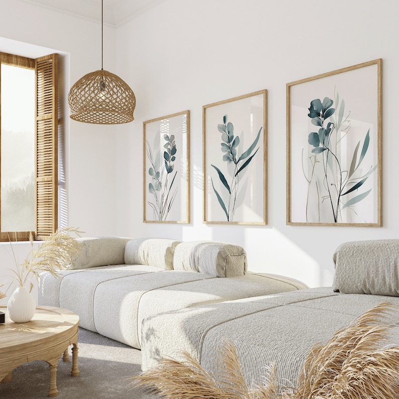 May include: Three framed botanical prints with a light background and blue-green foliage designs hang on a white wall above a white couch with two pillows. The couch is in a living room with a woven light fixture hanging from the ceiling.