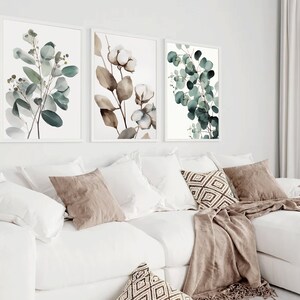 May include: Three framed botanical prints featuring watercolor illustrations of eucalyptus leaves and cotton blossoms. The prints are displayed on a white wall above a white couch with brown throw pillows and a brown blanket.