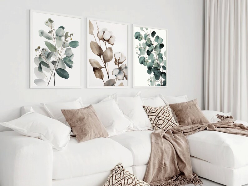 May include: Three framed botanical prints featuring watercolor illustrations of eucalyptus leaves and cotton blossoms. The prints are displayed on a white wall above a white couch with brown throw pillows and a brown blanket.