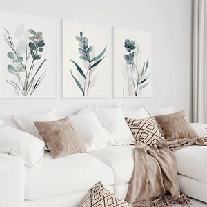 May include: Three framed watercolor prints of blue and gray floral designs on a white background. The prints are hanging on a white wall above a white couch with brown throw pillows and a brown blanket.