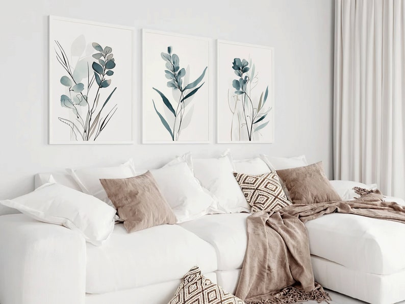 May include: Three framed watercolor prints of blue and gray floral designs on a white background. The prints are hanging on a white wall above a white couch with brown throw pillows and a brown blanket.