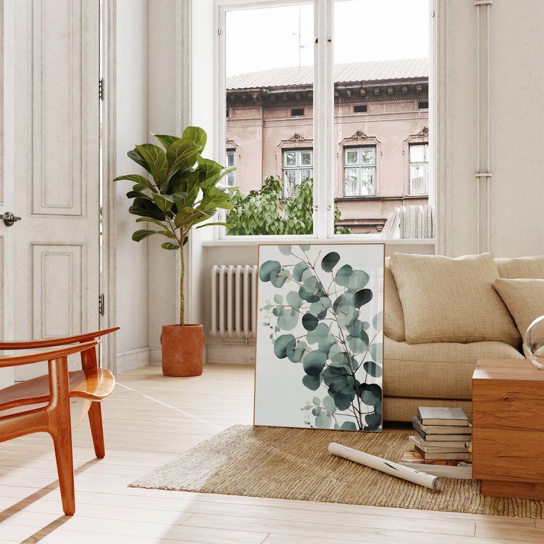 May include: A framed print of a watercolor painting of eucalyptus leaves in shades of green against a white background. The print is leaning against a beige couch in a living room setting.