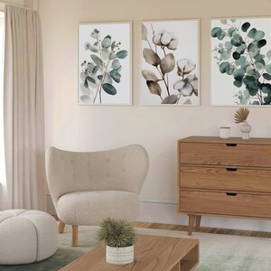 May include: A living room interior with a white armchair, a round ottoman, a wooden dresser with three drawers, and three framed botanical prints featuring green leaves and white flowers.