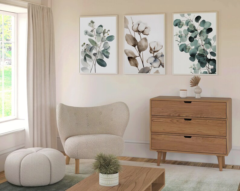 May include: A living room interior with a white armchair, a round ottoman, a wooden dresser with three drawers, and three framed botanical prints featuring green leaves and white flowers.