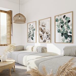 May include: Three framed botanical prints with watercolor illustrations of eucalyptus leaves and flowers in shades of green, brown, and white, hanging on a white wall above a white couch with pillows.
