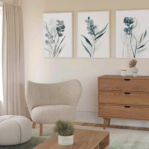 May include: Three framed watercolor prints of blue and green foliage hang on a light beige wall above a wooden dresser with three drawers. A white armchair, a white ottoman, and a potted green plant are in the foreground.