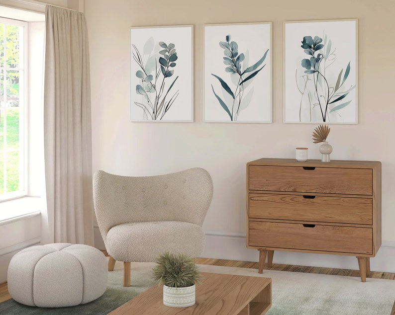May include: Three framed watercolor prints of blue and green foliage hang on a light beige wall above a wooden dresser with three drawers. A white armchair, a white ottoman, and a potted green plant are in the foreground.