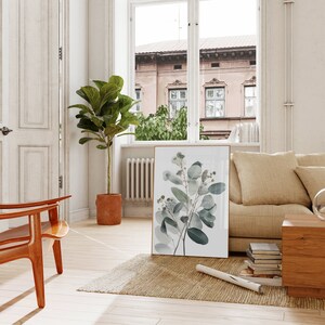 May include: A framed botanical print of a eucalyptus branch with green leaves and white flowers. The print is hanging on a white wall in a living room with a beige sofa, a wooden chair, and a potted plant.
