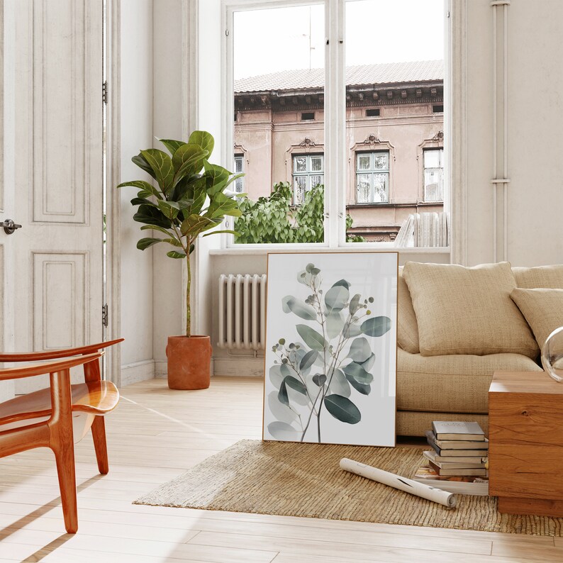 May include: A framed botanical print of a eucalyptus branch with green leaves and white flowers. The print is hanging on a white wall in a living room with a beige sofa, a wooden chair, and a potted plant.