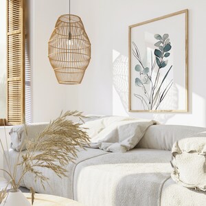 May include: A white couch with a light brown woven pendant lamp hanging above it. The couch has a white throw pillow and a white bolster pillow. A framed print with a blue and white floral design hangs on the wall behind the couch.