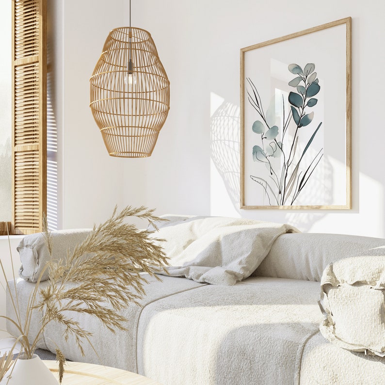 May include: A white couch with a light brown woven pendant lamp hanging above it. The couch has a white throw pillow and a white bolster pillow. A framed print with a blue and white floral design hangs on the wall behind the couch.