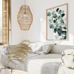 May include: A white couch with a light brown woven pendant lamp hanging above it. The couch has a few pillows and a throw blanket. A framed print of green leaves hangs on the wall behind the couch.