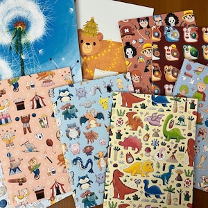 Op de afbeelding: Een verzameling kleurrijke notitieboekjes met diverse illustraties. Ontwerpen omvatten een paardenbloem, een beer met een kerstboom, stripfiguren, circustafereel, dinosaurussen en fantasie wezens. De notitieboekjes liggen op een houten oppervlak.